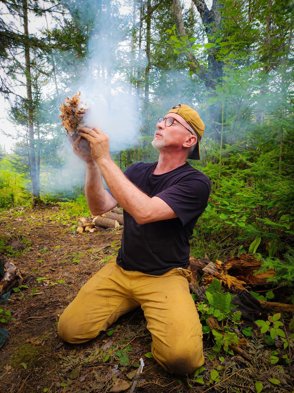 Man in a forest setting holding a lit piece of wood with smoke