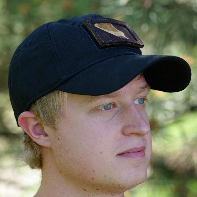A man wearing a black baseball cap with a camo patch attached to the front.