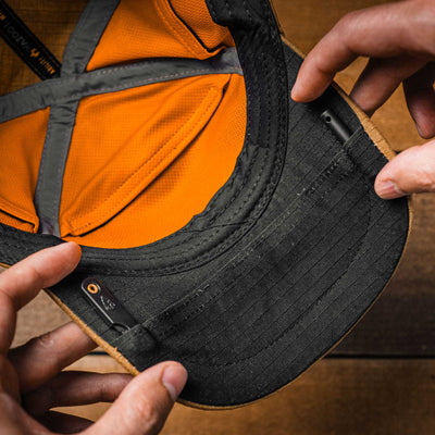 Close-up of a black cap with hands putting items into pockets on the brim of the hat.