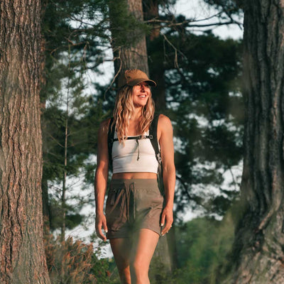 A woman standing in the woods, wearing a coyote brown baseball cap, white tank top, and green shorts.