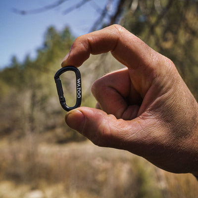 Wazoo micro carabiner shown held between two fingers in the outdoors highlighting its tiny compact size