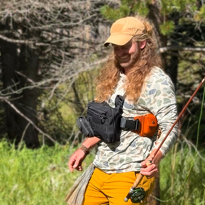 A man wearing a coyote brown baseball cap, holding a fishing pole, in the woods.